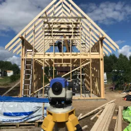 Wznoszenie szkieletu domu z drewnianych belek, widoczny niwelator optyczny na statywie, pracownik na wysokości, drabina oparta o konstrukcję, błękitne niebo z chmurami w tle.