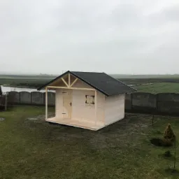 Surowa drewniana konstrukcja altany z czarnym dachem, widoczna z zewnątrz na tle pochmurnego nieba i zielonego pola, z elementami ogrodu w pierwszym planie.