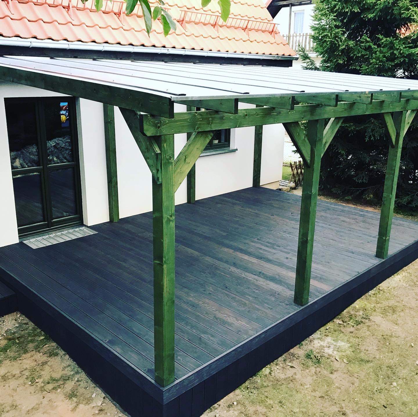 Zadaszony taras z ciemnego drewna, zielone drewniane słupy, widok z zewnątrz, częściowo zacieniony, obok domu z czerwoną dachówką.