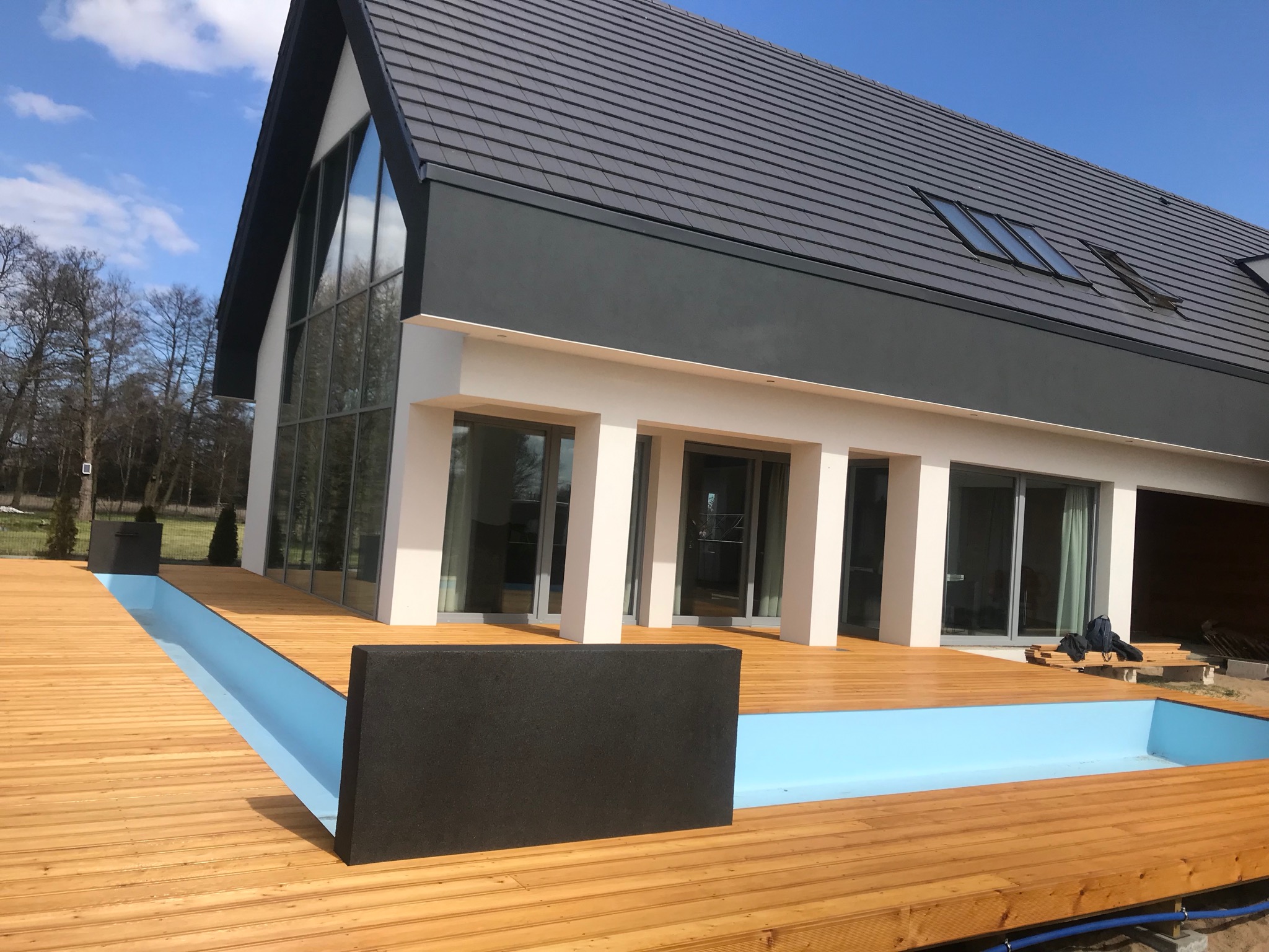 Nowoczesny dom z dużymi oknami i tarasem z drewna z wbudowanym basenem o błękitnym dnie. W tle drzewa i błękitne niebo.