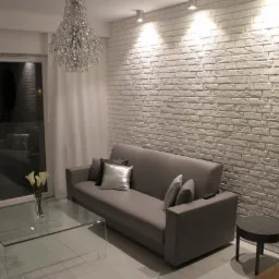 Jasne wnętrze pokoju dziennego z sofą, szklanym stolikiem kawowym i ścianą z białej cegły dekoracyjnej podświetloną reflektorami.