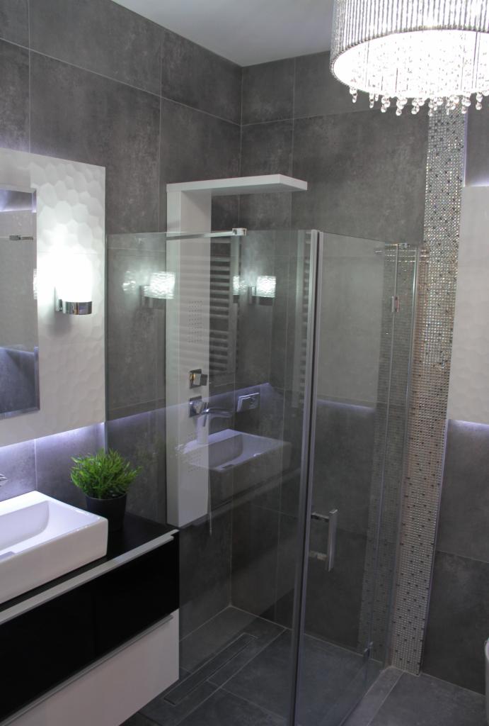 Nowoczesna łazienka z szarymi płytkami, szklaną kabiną prysznicową, umywalką nablatową, dekoracyjnym oświetleniem LED i kryształowym żyrandolem.