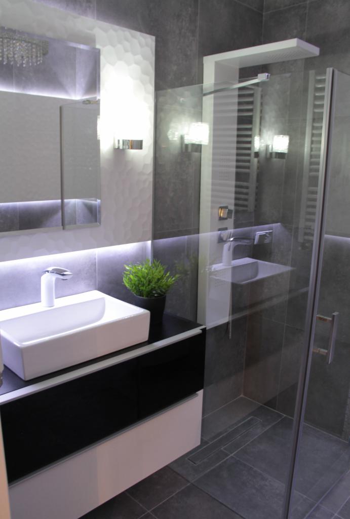 Nowoczesna łazienka z szarą glazurą, białą umywalką nablatową na czarno-białej szafce, podświetlanym lustrem i kabiną prysznicową ze szklanymi drzwiami.