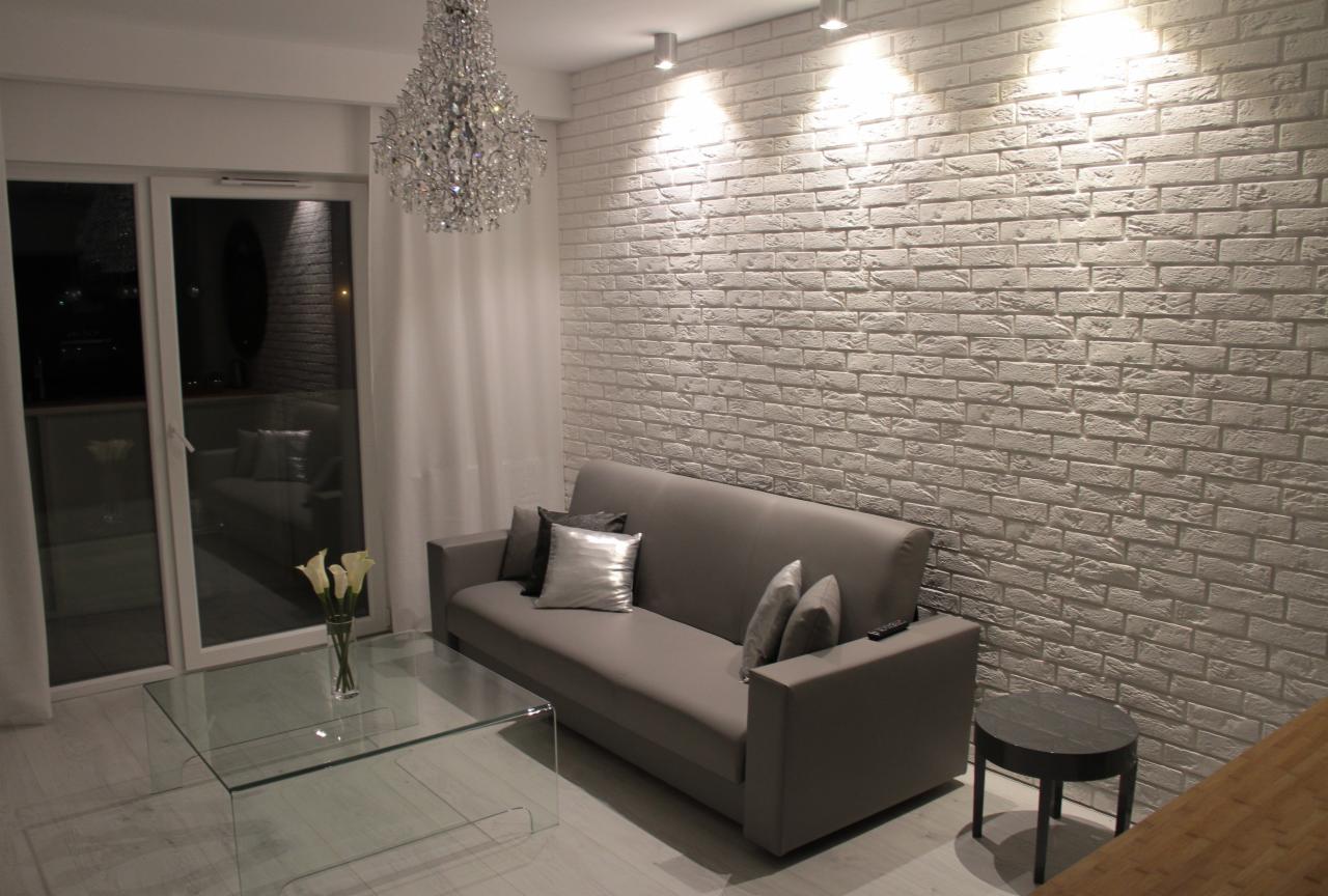 Jasne wnętrze pokoju dziennego z sofą, szklanym stolikiem kawowym i ścianą z białej cegły dekoracyjnej podświetloną reflektorami.