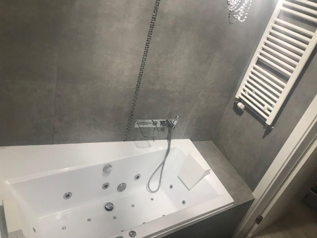Nowoczesna łazienka z szarą płytką, białą wanną z hydromasażem i grzejnikiem drabinkowym, widoczny fragment kryształowego żyrandola.