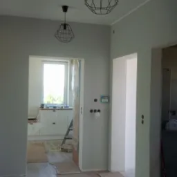 Wnętrze mieszkania w trakcie prac wykończeniowych: świeżo pomalowane szare ściany, zabezpieczona podłoga z paneli imitujących drewno, widoczne czarne lampy druciane i fragment kuchni w tle.