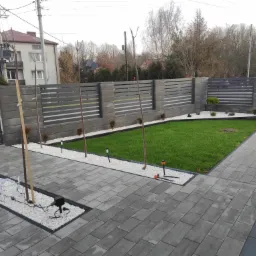 Nowoczesny ogród z szarą kostką brukową, ogrodzeniem z poziomych paneli i trawnikiem otoczonym białymi kamieniami, widoczne młode drzewka i ozdobna iluminacja w kształcie renifera.