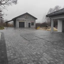 Ułożona szara kostka brukowa na podjeździe, z widocznymi ciemniejszymi kwadratami, obok budynków w trakcie budowy, w pochmurny dzień.