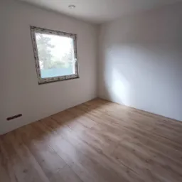 Pusty pokój po odświeżeniu z panelami podłogowymi w odcieniach drewna, świeżo pomalowanymi ścianami na biało i nowym oknem z widokiem na drzewa.