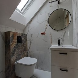 Usługi remontowo - budowlane Zbigniew Rzepka - Nowoczesna łazienka na poddaszu z marmurowymi płytkami, owalnym lustrem i wiszącą toaletą obudowaną płytkami imitującymi kamień.