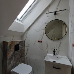 Usługi remontowo - budowlane Zbigniew Rzepka - Nowoczesna łazienka na poddaszu z oknem dachowym, białe płytki imitujące marmur z delikatnym złotym żyłkowaniem, okrągłe lustro z czarną ramą i minimalistyczna lampa LED nad umywalką, wisząca...