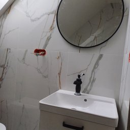 Usługi remontowo - budowlane Zbigniew Rzepka - Wykończona łazienka z umywalką na szafce, czarną baterią retro, okrągłym lustrem w czarnej ramie i białymi płytkami imitującymi marmur z delikatnymi złotymi żyłami.