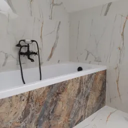 Biała wanna z czarną armaturą w łazience wykończonej płytkami imitującymi marmur i kamień, z kontrastującą okładziną w brązowo-szarej kolorystyce na obudowie wanny.