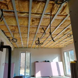 Wnętrze w Łasku w trakcie prac: widok na sufit z zamontowaną konstrukcją metalową, ocieplony wełną mineralną, z wystającymi kablami elektrycznymi, w tle ściany i okna.