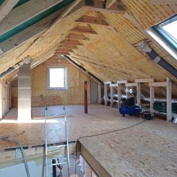 Usługi remontowo - budowlane Zbigniew Rzepka - Poddasze w trakcie adaptacji: ocieplona skośna ściana z widoczną izolacją, drewniana konstrukcja dachu, okno dachowe, kompresor i podłoga z płyt OSB.