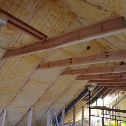 Usługi remontowo - budowlane Zbigniew Rzepka - Żółta izolacja z wełny mineralnej zamontowana na poddaszu, widoczne drewniane krokwie, sznurki mocujące, rury wentylacyjne i metalowa drabina.