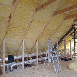 Usługi remontowo - budowlane Zbigniew Rzepka - Poddasze z widoczną izolacją z wełny mineralnej, drewnianą konstrukcją nośną, srebrnymi rurami wentylacyjnymi, aluminiową drabiną i narzędziem na podłodze z płyt OSB.