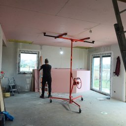 Usługi remontowo - budowlane Zbigniew Rzepka - Montaż płyt kartonowo-gipsowych na suficie, widoczny pracownik z podnośnikiem, częściowo ocieplone poddasze, surowe ściany i okna.