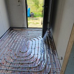 Usługi remontowo - budowlane Zbigniew Rzepka - Rozłożone rury ogrzewania podłogowego na folii z czerwoną kratką w surowym pomieszczeniu z otwartymi drzwiami na zewnątrz.