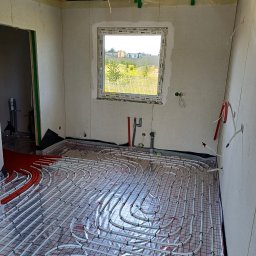 Usługi remontowo - budowlane Zbigniew Rzepka - Instalacja ogrzewania podłogowego w surowym pomieszczeniu z widocznymi rurami grzewczymi, izolacją i nieotynkowanymi ścianami z widocznymi przewodami elektrycznymi, przez okno widok na zabudowania.