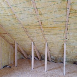 Usługi remontowo - budowlane Zbigniew Rzepka - Poddasze w trakcie ocieplania wełną mineralną, widoczne drewniane krokwie i zielone sznurki zabezpieczające izolację, podłoga wyłożona płytą OSB, fragment instalacji elektrycznej.