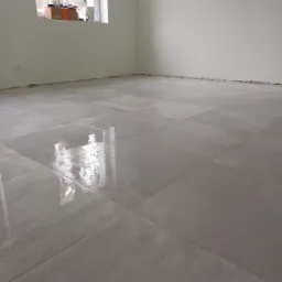 Świeżo ułożone, mokre, szare płytki podłogowe w pustym pokoju, widoczne ślady po fugowaniu i okna w tle.