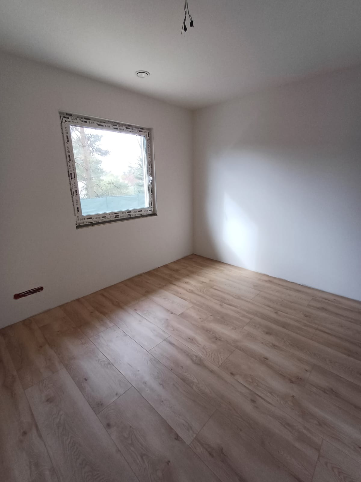 Pusty pokój po odświeżeniu z panelami podłogowymi w odcieniach drewna, świeżo pomalowanymi ścianami na biało i nowym oknem z widokiem na drzewa.