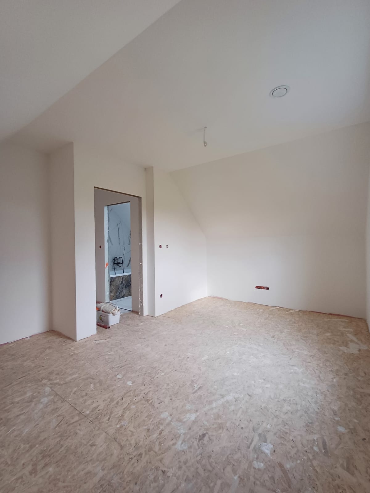 Poddasze w trakcie prac wykończeniowych: świeżo pomalowane ściany na biało, surowa podłoga z płyt OSB, widoczne gniazdka elektryczne, otwór drzwiowy prowadzący do łazienki z marmurowymi płytkami.