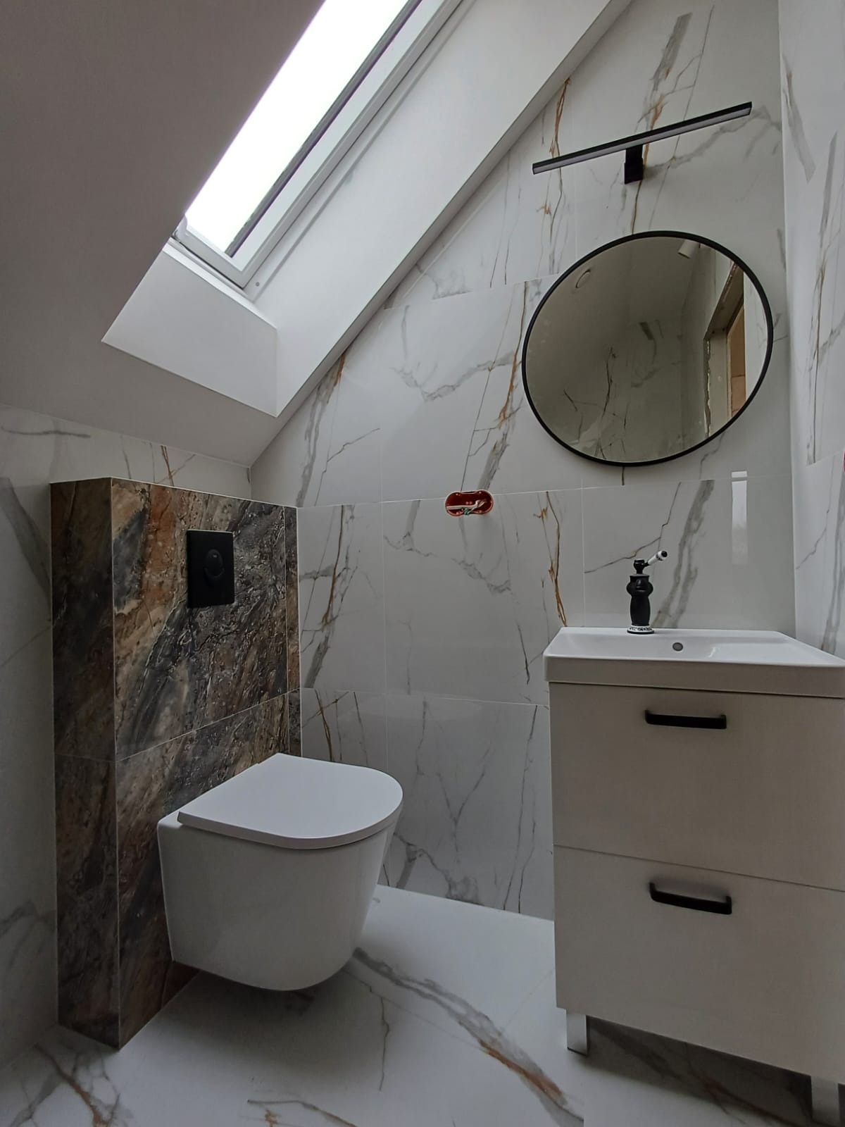 Nowoczesna łazienka na poddaszu z marmurowymi płytkami, owalnym lustrem i wiszącą toaletą obudowaną płytkami imitującymi kamień.