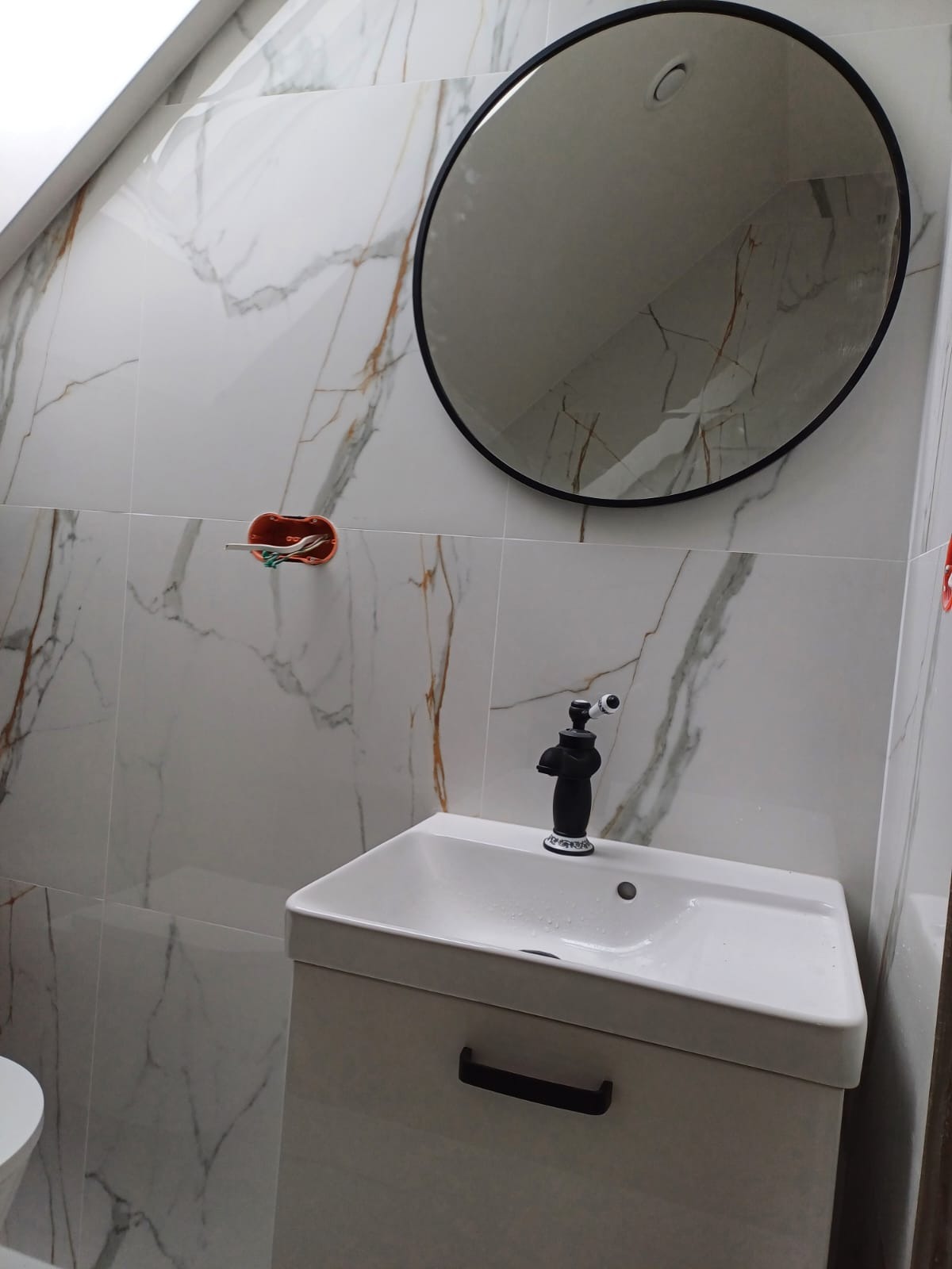 Wykończona łazienka z umywalką na szafce, czarną baterią retro, okrągłym lustrem w czarnej ramie i białymi płytkami imitującymi marmur z delikatnymi złotymi żyłami.