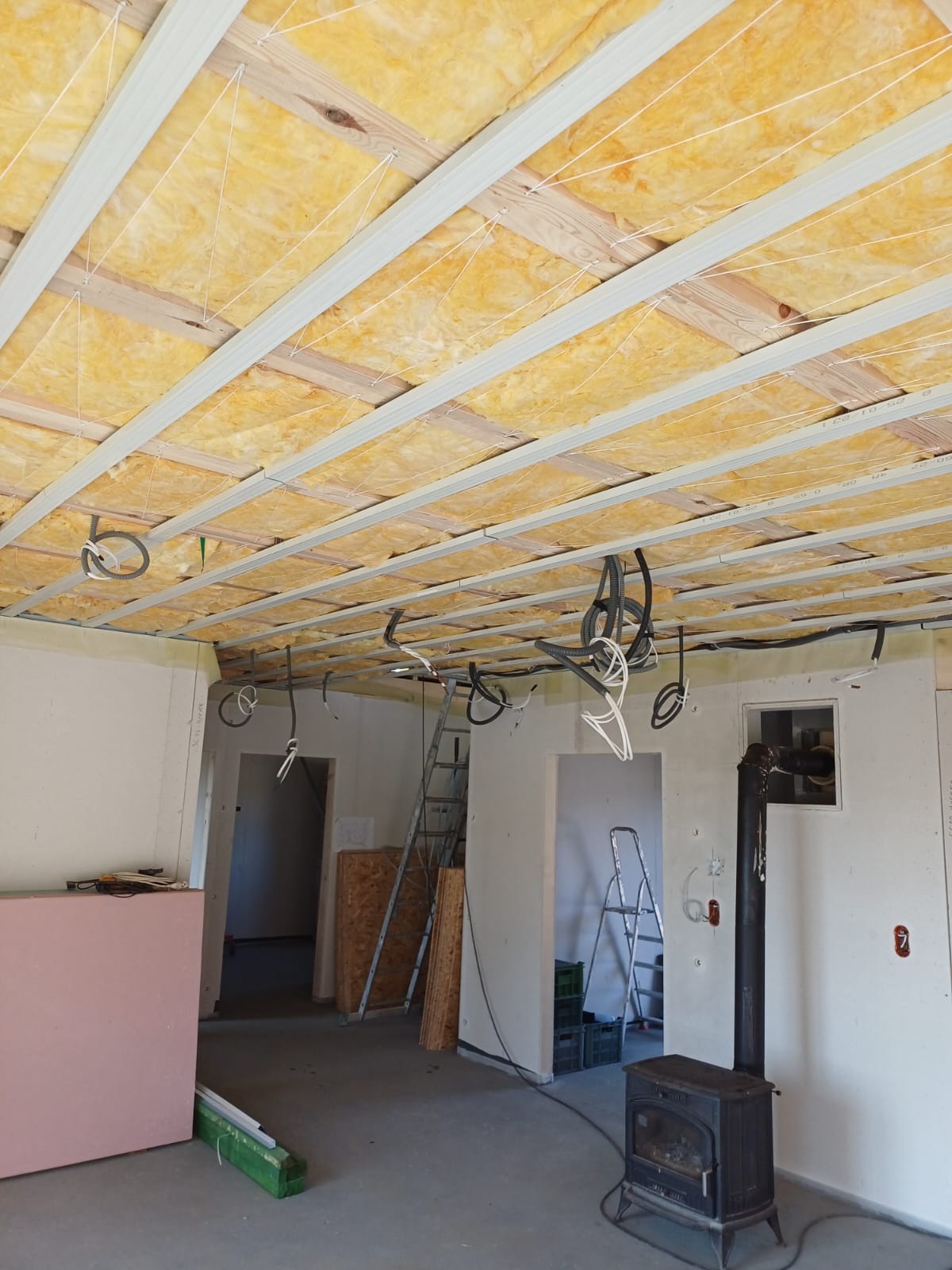 Poddasze w trakcie ocieplania wełną mineralną, widoczne profile metalowe, drewniane belki, zwisające przewody elektryczne i drabina oparta o ścianę w surowym wnętrzu.