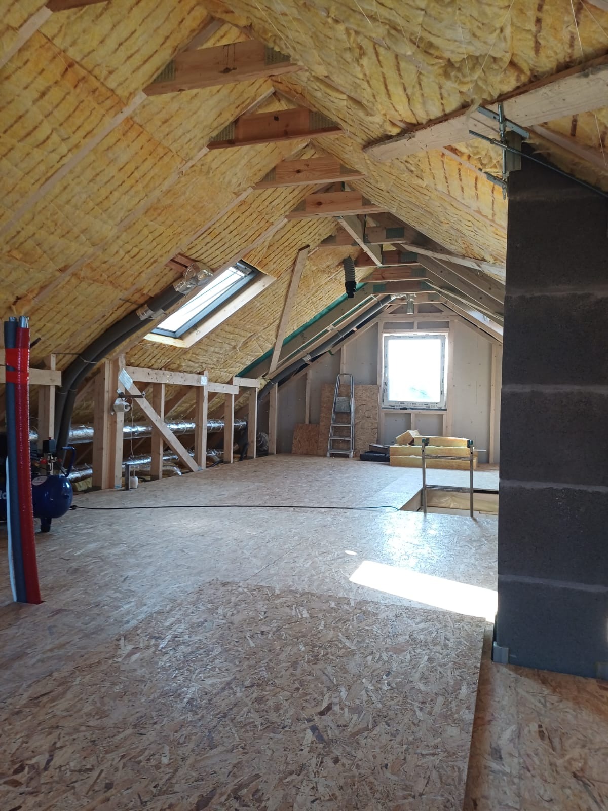 Poddasze z ociepleniem z wełny mineralnej, widoczne okno dachowe, konstrukcja drewniana, płyta OSB na podłodze oraz kompresor powietrza przy ścianie.