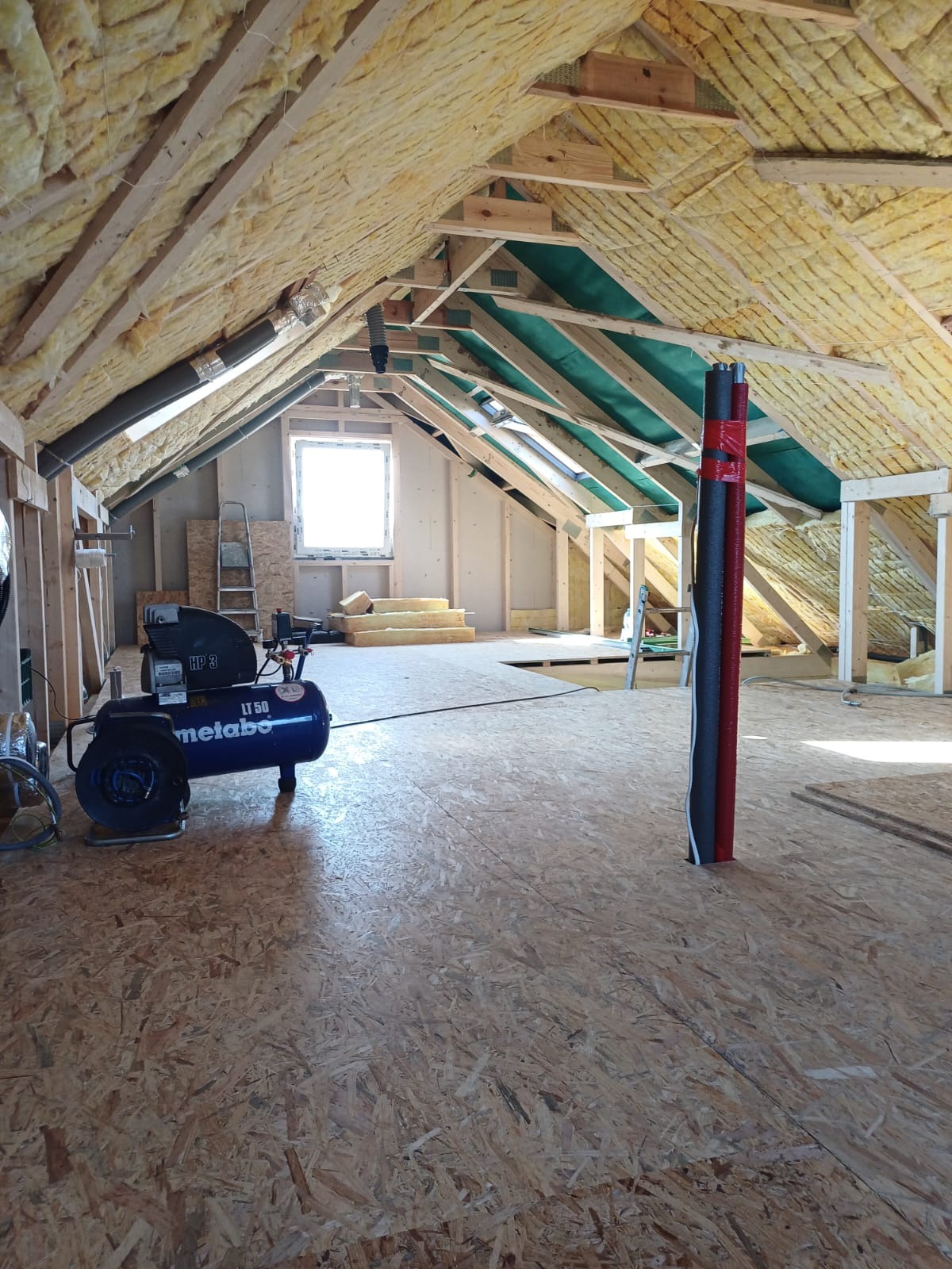 Poddasze w trakcie adaptacji: widoczna izolacja z wełny mineralnej między krokwiami, podłoga z płyt OSB, kompresor Metabo LT 50 i pionowe rury owinięte czerwoną taśmą.