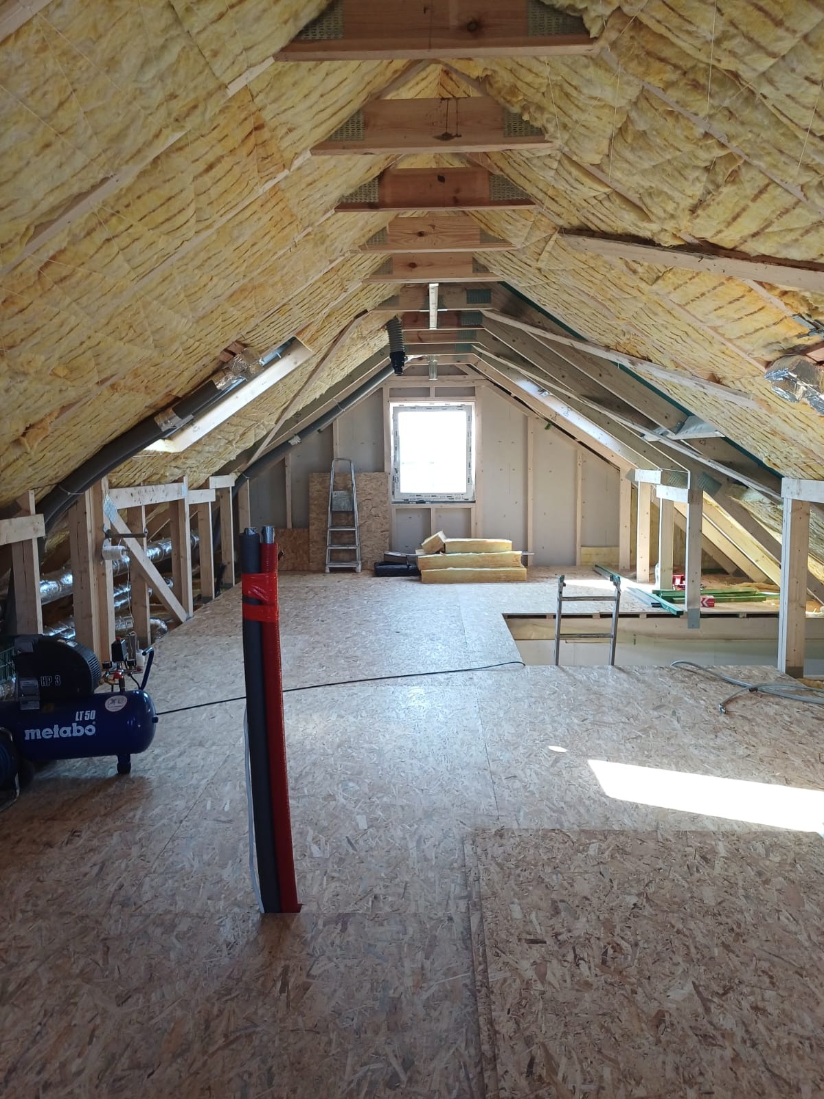 Poddasze w trakcie prac wykończeniowych: ocieplone skosy dachu, drewniana konstrukcja, częściowo położona płyta podłogowa OSB, kompresor Metabo i rury wentylacyjne.