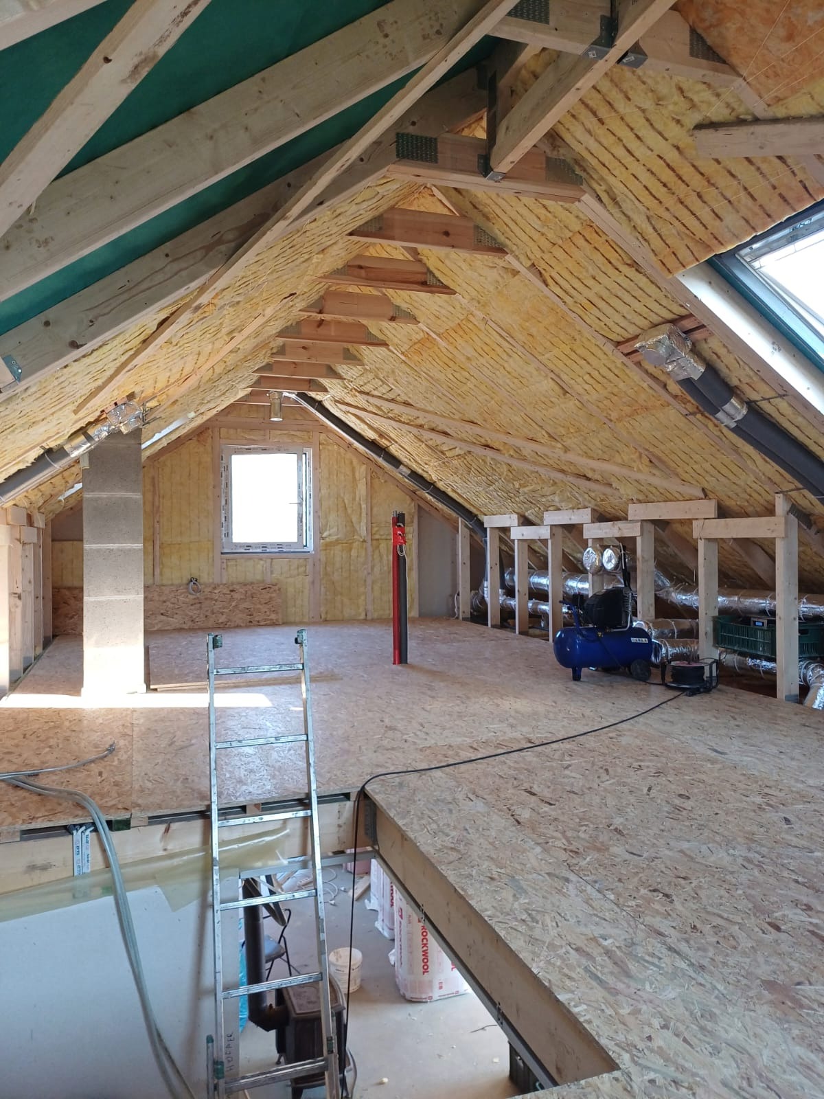 Poddasze w trakcie adaptacji: ocieplona skośna ściana z widoczną izolacją, drewniana konstrukcja dachu, okno dachowe, kompresor i podłoga z płyt OSB.