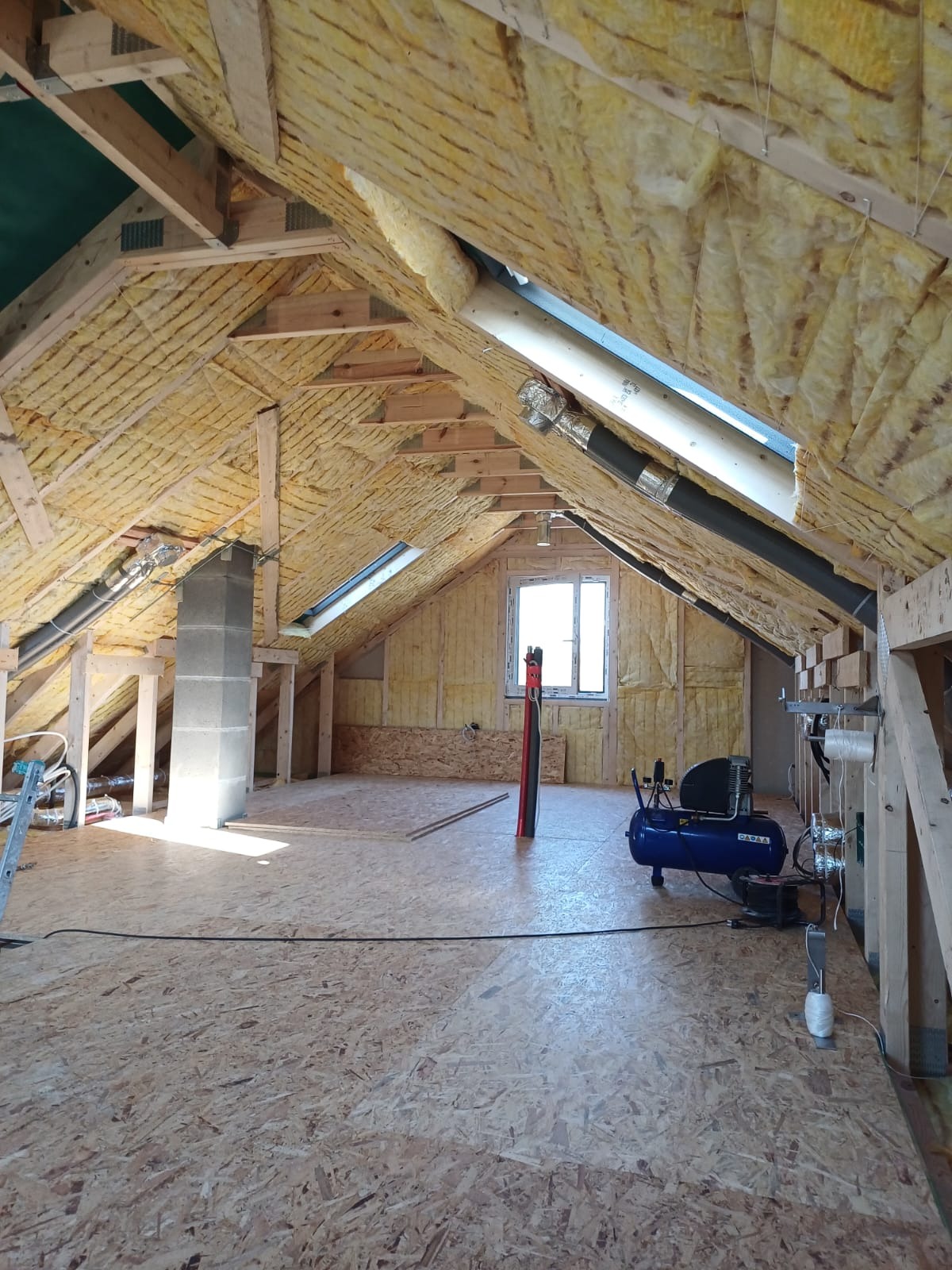 Poddasze w trakcie ocieplania wełną mineralną, widoczne drewniane krokwie, okno dachowe, komin, niebieski kompresor i częściowo ułożona podłoga z płyt OSB.