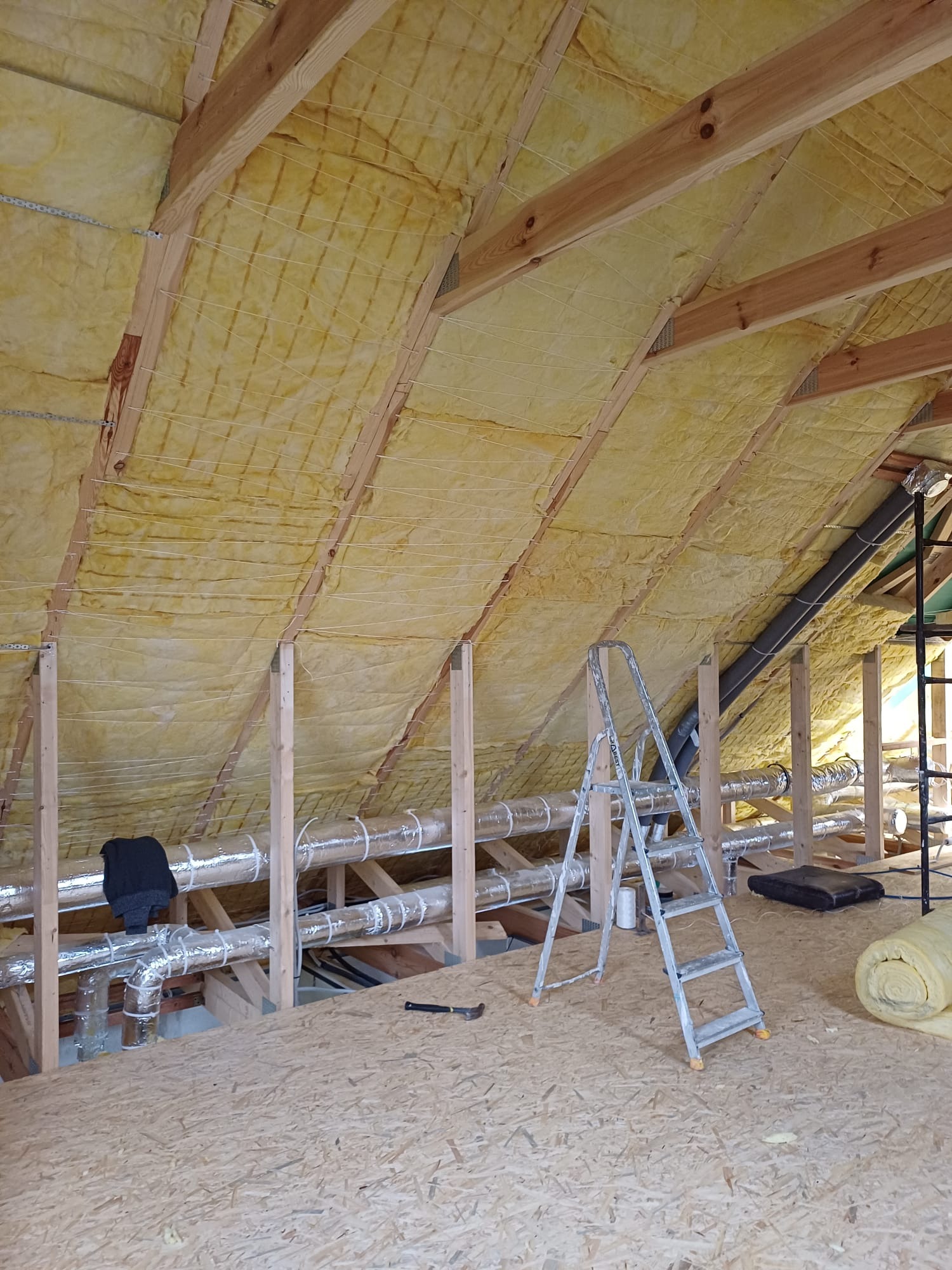 Poddasze z widoczną izolacją z wełny mineralnej, drewnianą konstrukcją nośną, srebrnymi rurami wentylacyjnymi, aluminiową drabiną i narzędziem na podłodze z płyt OSB.