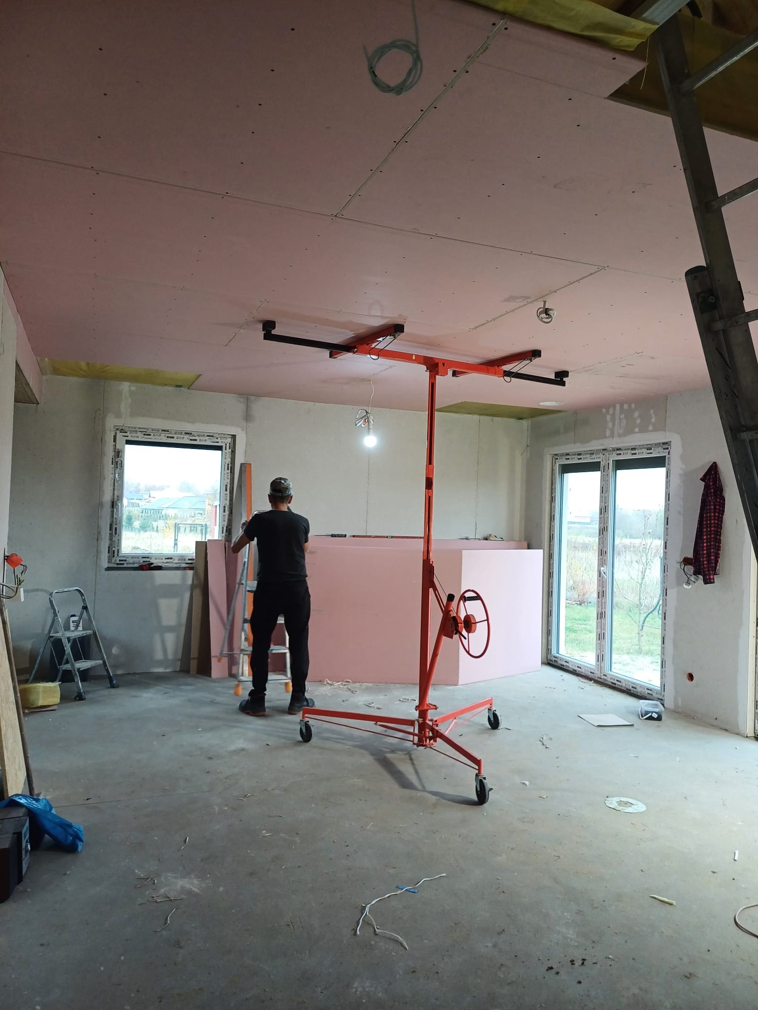 Montaż płyt kartonowo-gipsowych na suficie, widoczny pracownik z podnośnikiem, częściowo ocieplone poddasze, surowe ściany i okna.