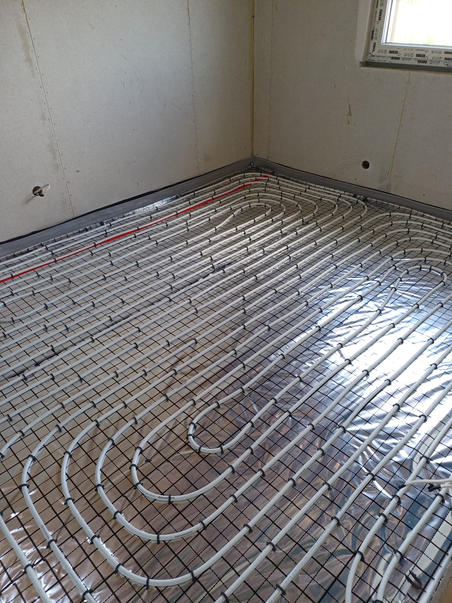 Rozłożona instalacja ogrzewania podłogowego na folii izolacyjnej z widocznymi rurkami w kolorze białym i elementami mocującymi w nowo wykończonym pomieszczeniu.