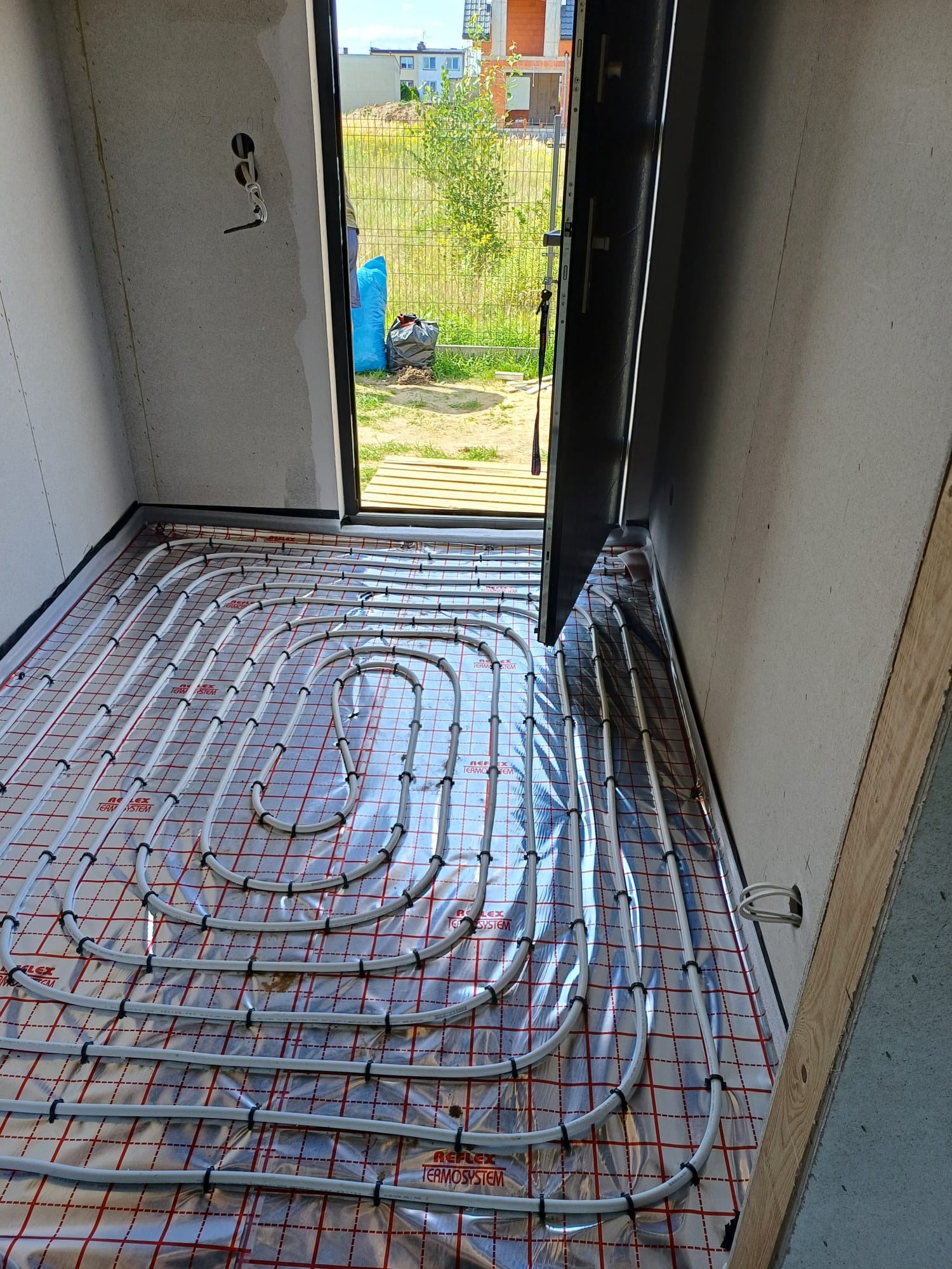 Rozłożone rury ogrzewania podłogowego na folii z czerwoną kratką w surowym pomieszczeniu z otwartymi drzwiami na zewnątrz.