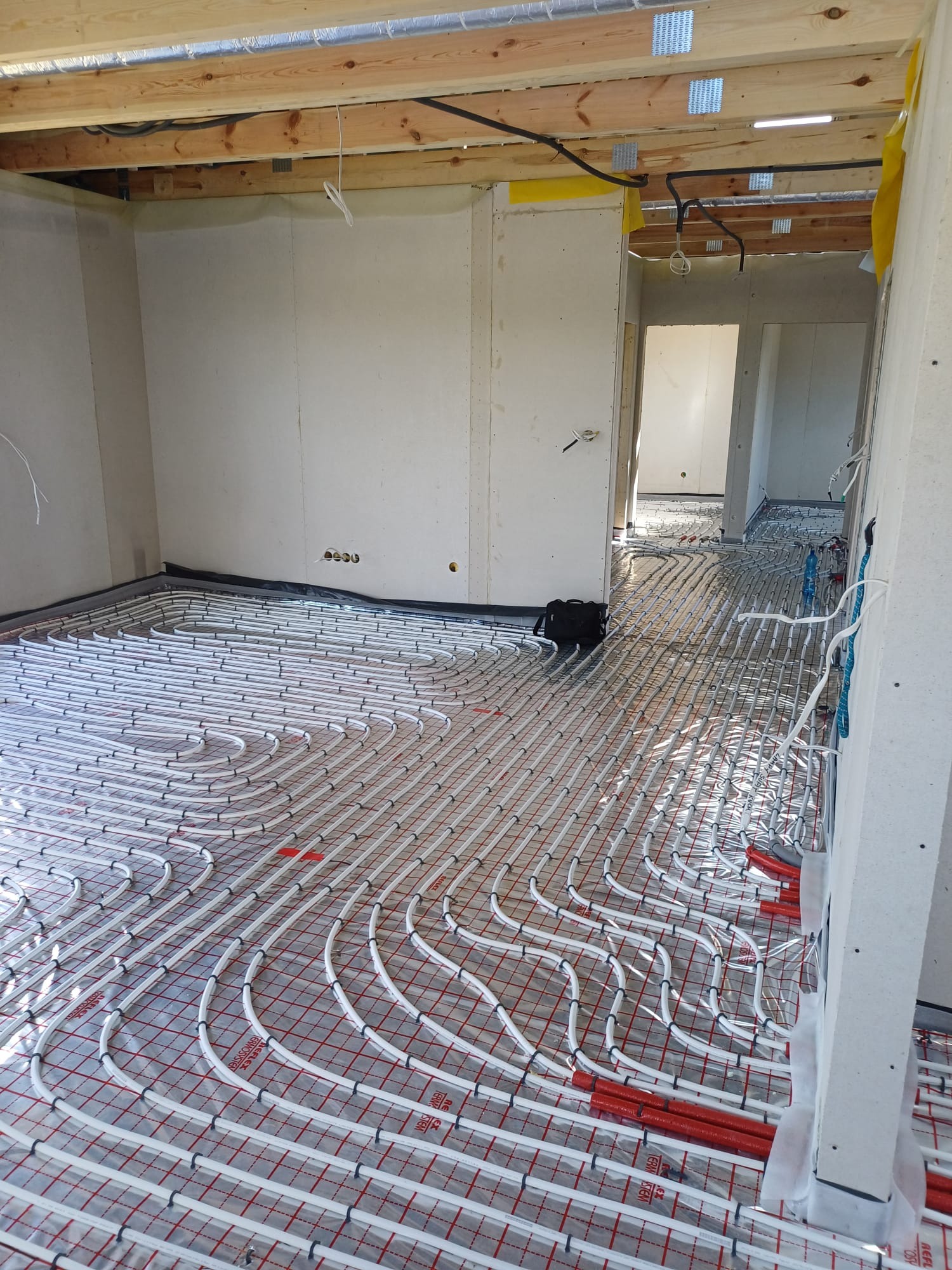 Rozłożona instalacja ogrzewania podłogowego na folii z siatką, widoczne rury grzewcze w kolorze białym i czerwonym, surowy stan ścian i drewniany strop.