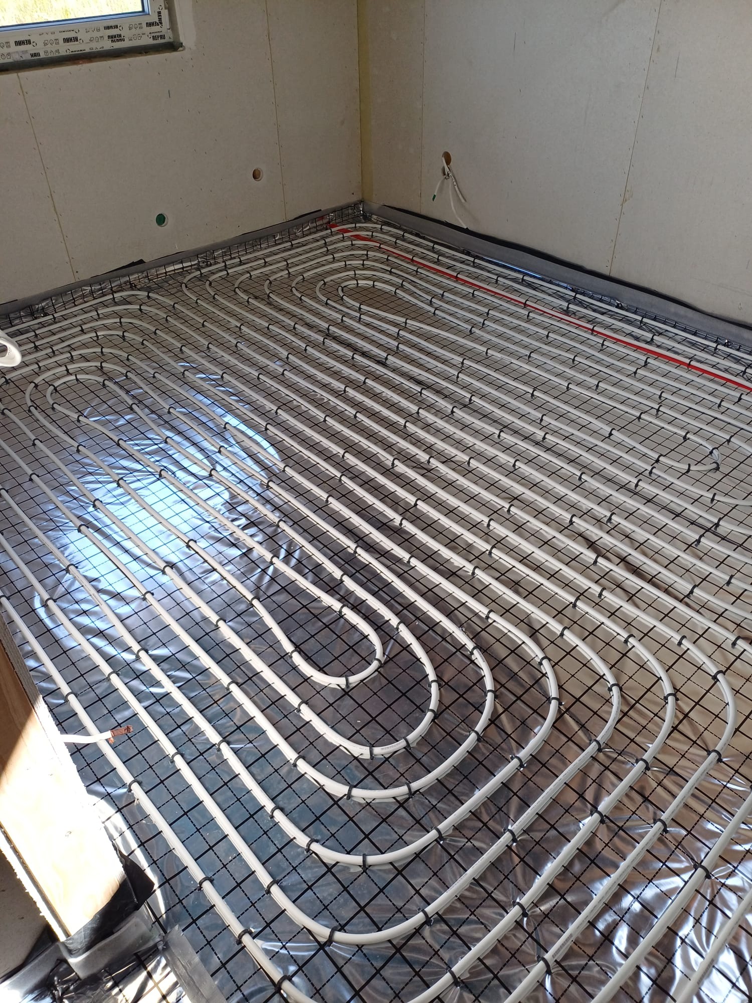 Rozłożona instalacja ogrzewania podłogowego z białych rurek na srebrnej folii izolacyjnej, widoczne czarne uchwyty mocujące i fragment czerwonej rurki, surowa ściana w tle.
