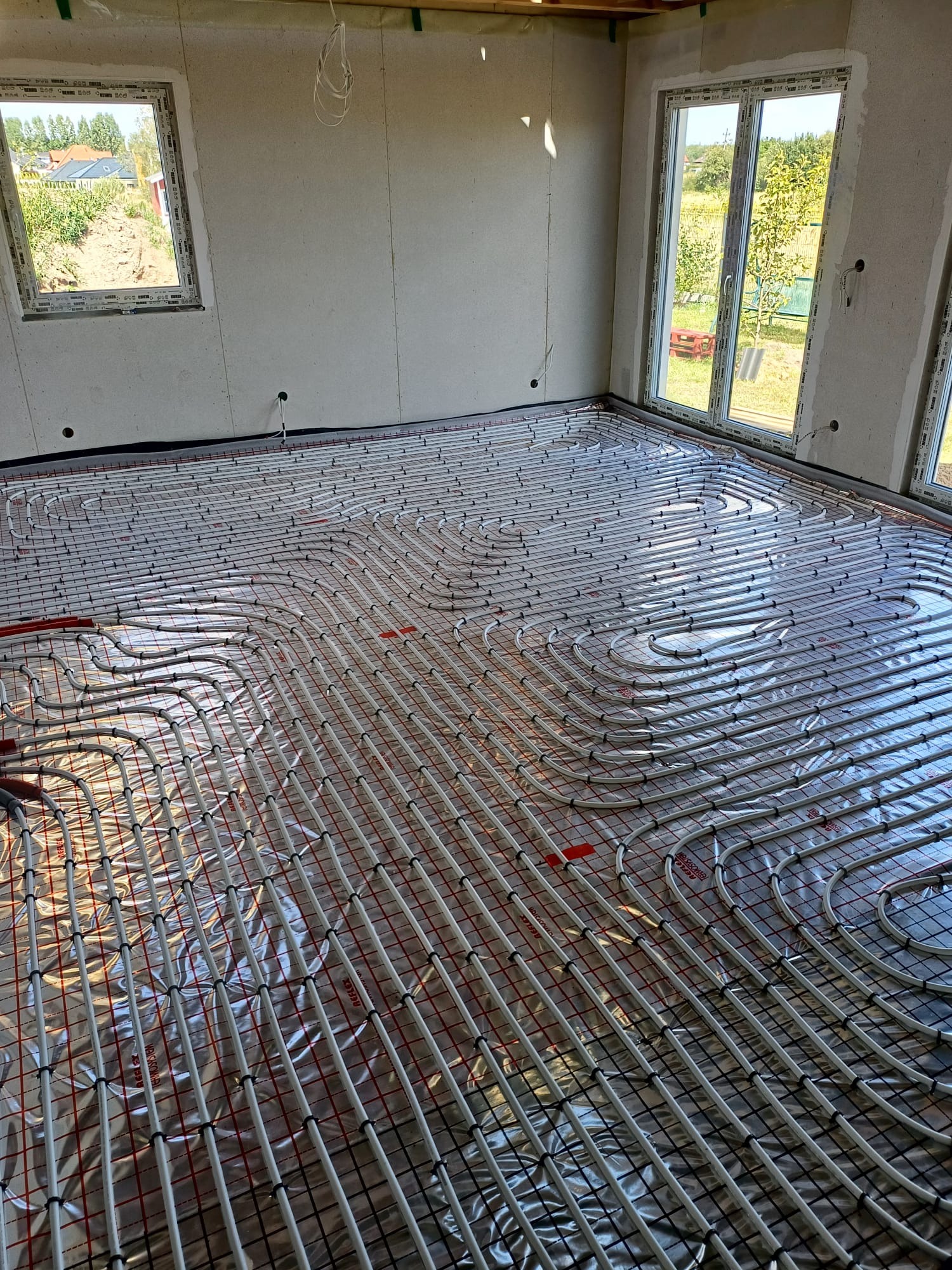 Rozłożona instalacja ogrzewania podłogowego na folii termoizolacyjnej w surowym pomieszczeniu z oknami i widokiem na zieleń na zewnątrz.
