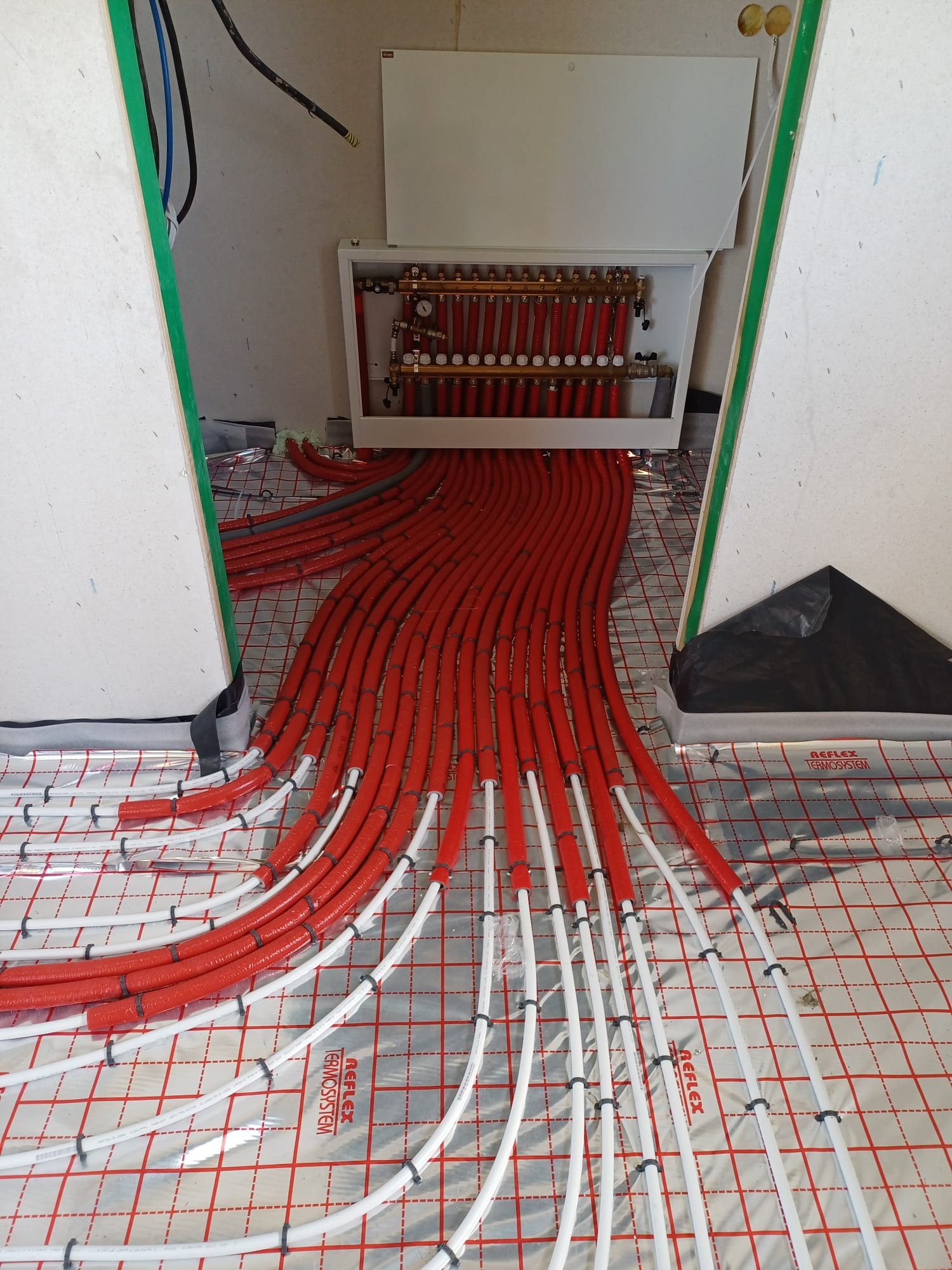 Rozdzielacz ogrzewania podłogowego z czerwonymi rurami grzewczymi na srebrnej folii izolacyjnej z nadrukiem siatki, widoczne elementy instalacji i przygotowane podłoże.