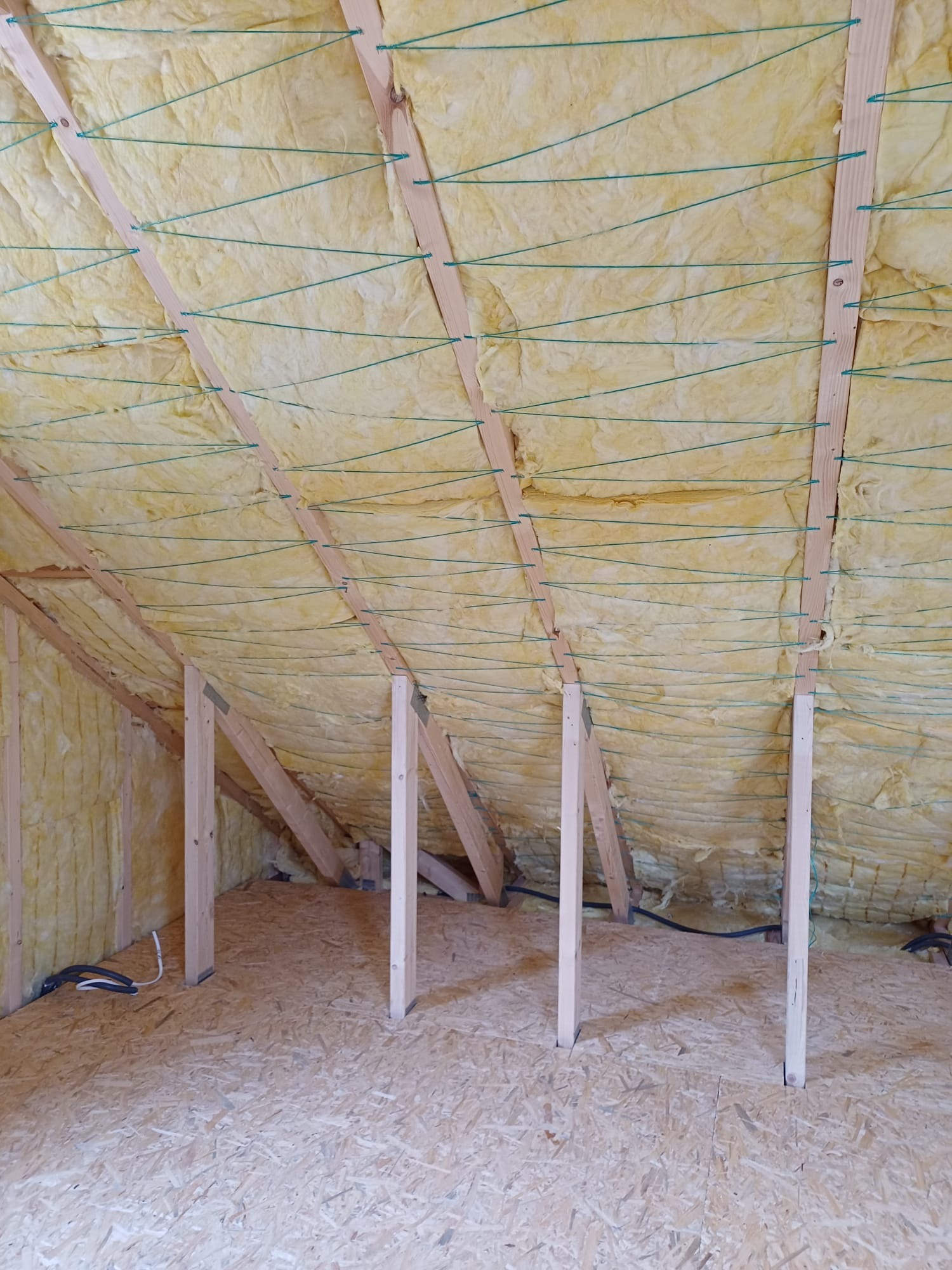 Poddasze w trakcie ocieplania wełną mineralną, widoczne drewniane krokwie i zielone sznurki zabezpieczające izolację, podłoga wyłożona płytą OSB, fragment instalacji elektrycznej.