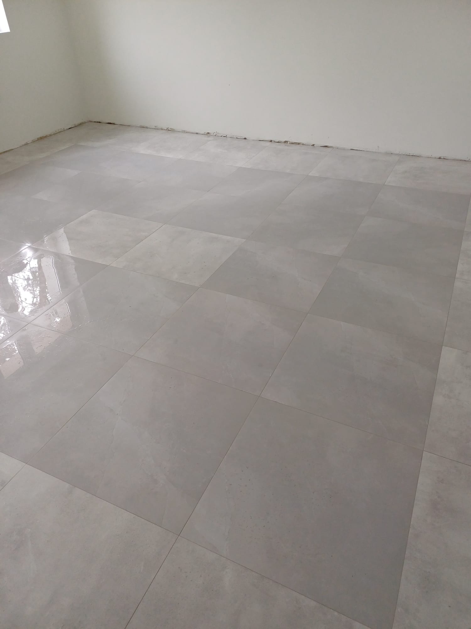 Świeżo ułożone, duże, matowe płytki podłogowe w odcieniach szarości, widoczne ślady po kleju na ścianie, przygotowanie do dalszych prac wykończeniowych.