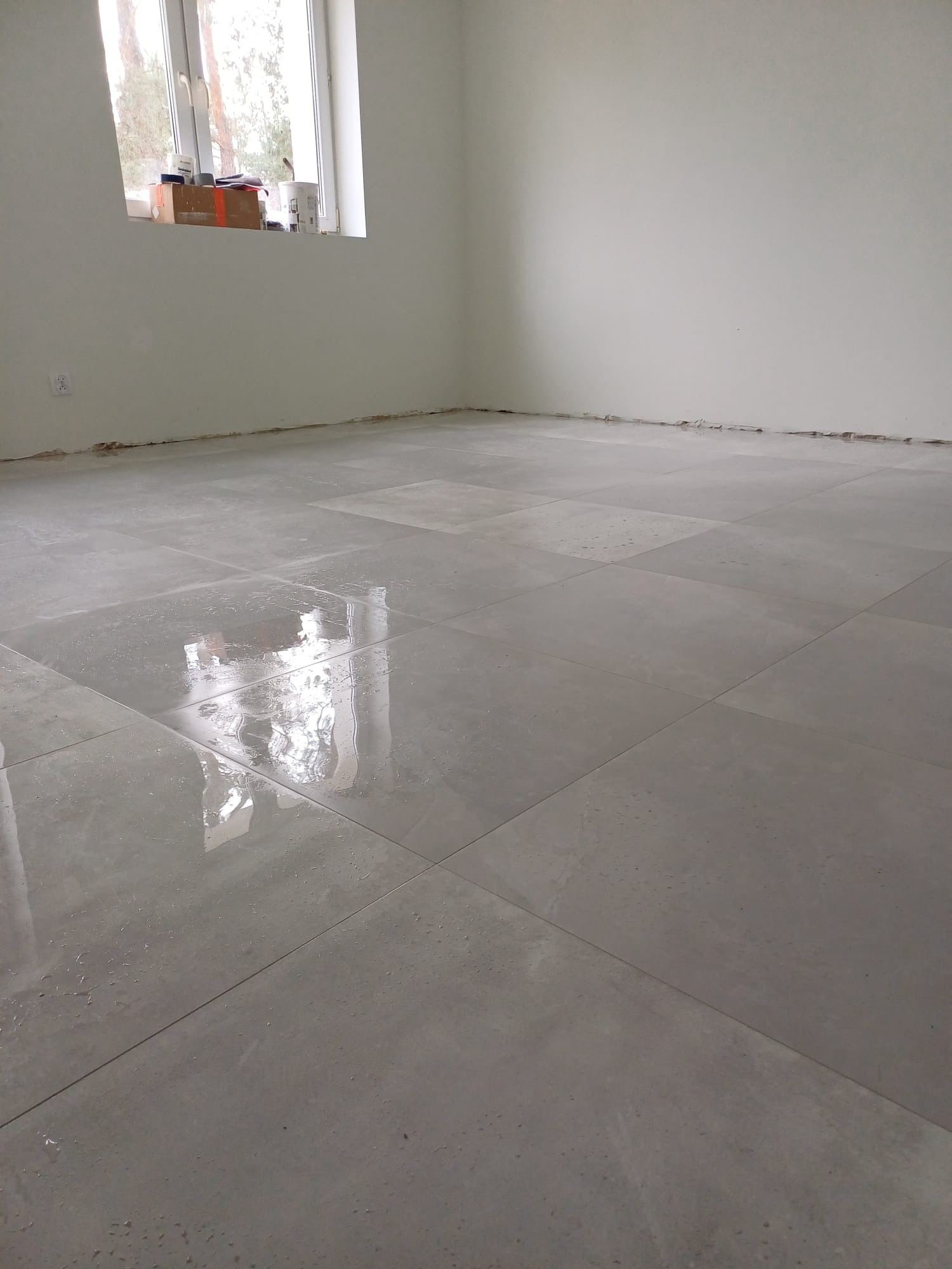Świeżo ułożone, mokre, szare płytki podłogowe w pustym pokoju, widoczne ślady po fugowaniu i okna w tle.