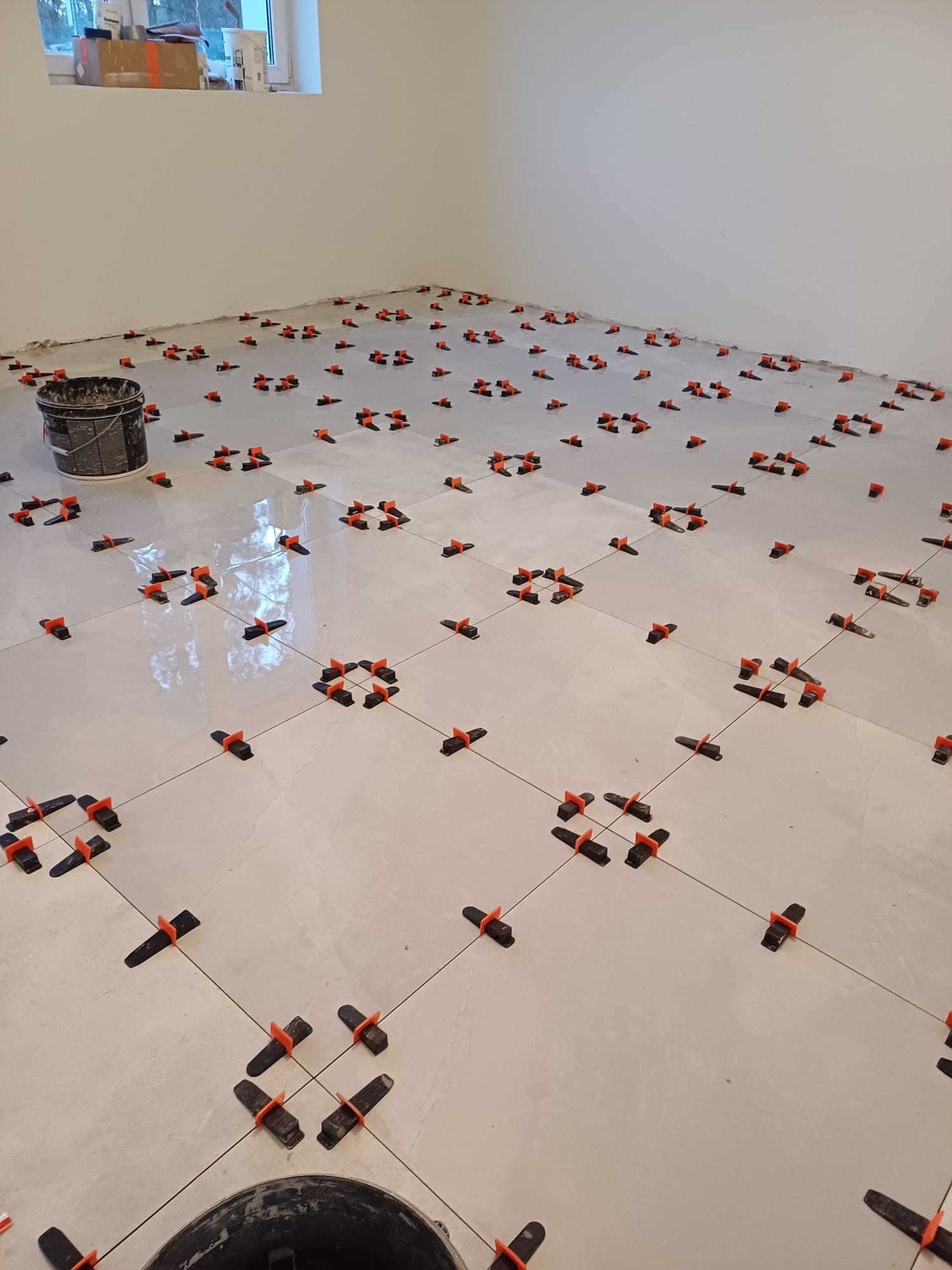 Jasne płytki podłogowe w trakcie układania, z systemem poziomowania płytek (klipsy i kliny) oraz wiadro z zaprawą w rogu pomieszczenia.