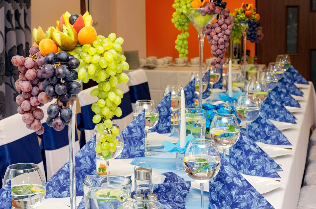 Uroczysty stół z dekoracjami z owoców, winogron w wysokich kielichach, niebieskie serwetki w kwiatowy wzór, woda z cytryną w szklankach, białe świece i dekoracyjne niebieskie wstążki.
