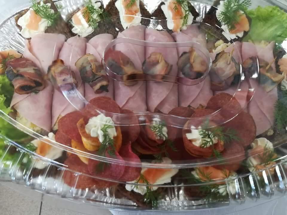 Taca z różnorodnymi przekąskami: roladki z szynki, koreczki z salami i kremowym serem, kanapeczki z łososiem i koperkiem, całość pod przezroczystą pokrywą.